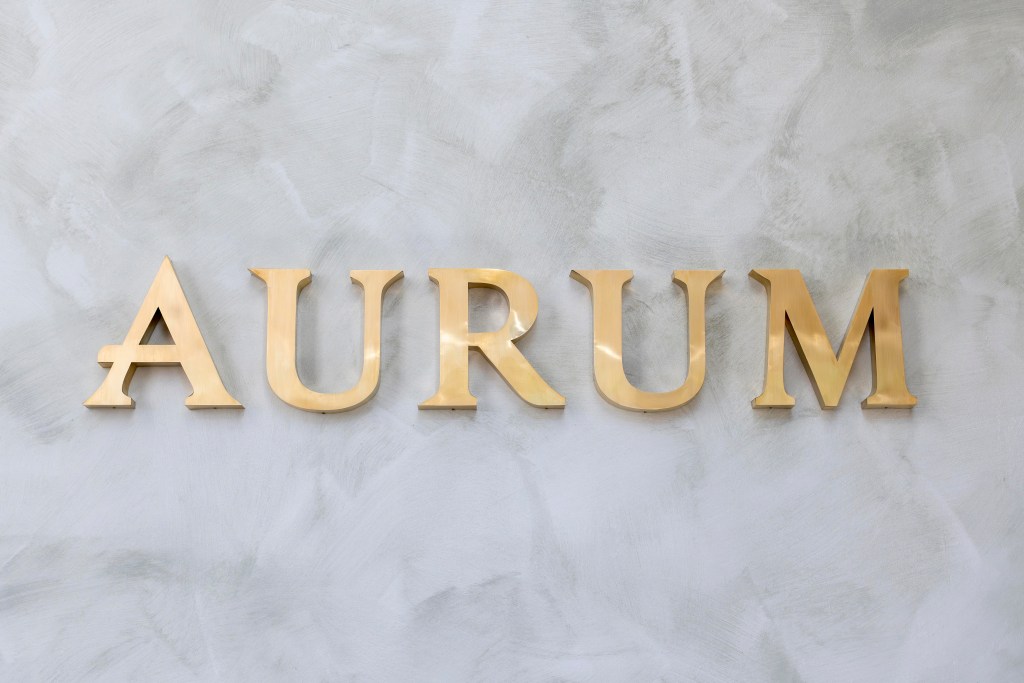 Logotipo dorado que dice 'AURUM' sobre un fondo de pared texturizada en tonos grises.